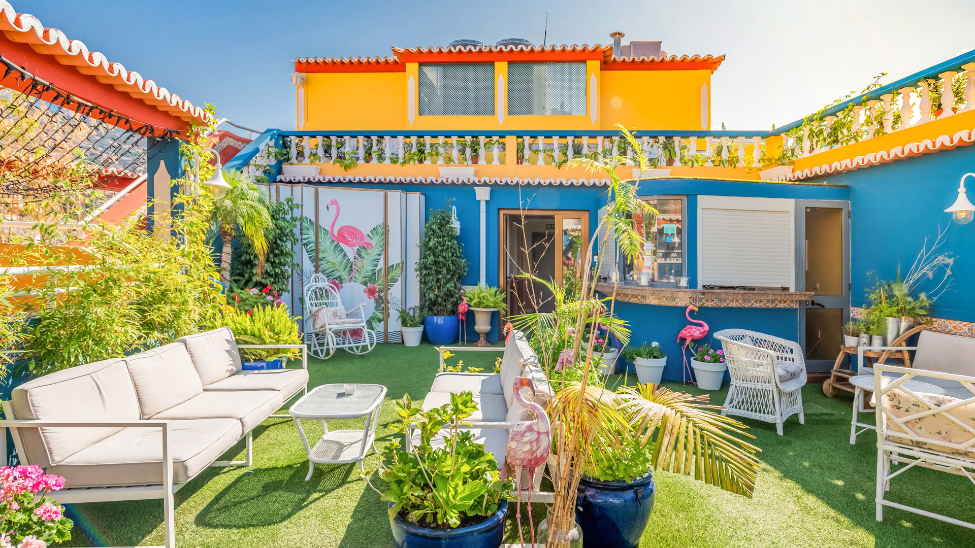 Terraço com plantas, sofás e decoração junto a prédio amarelo.