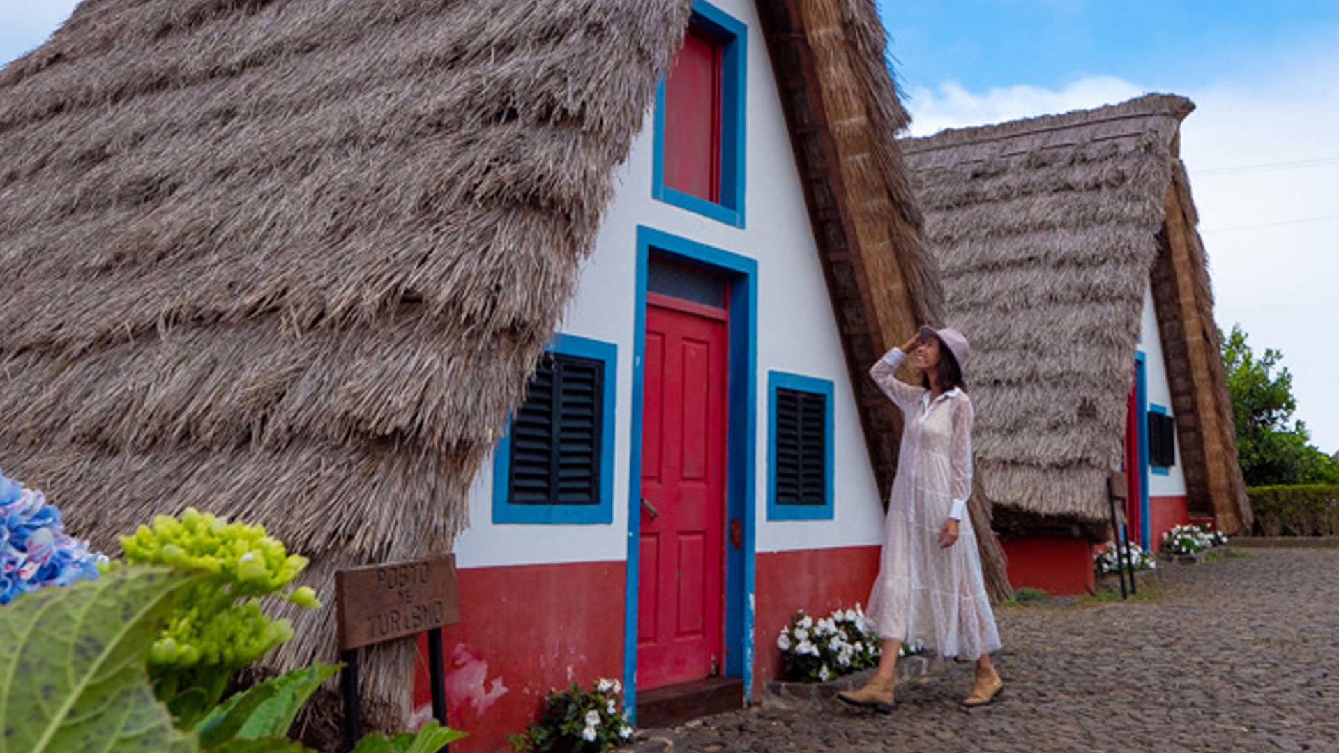 Woman in white dress and hat near house with thatched roof.