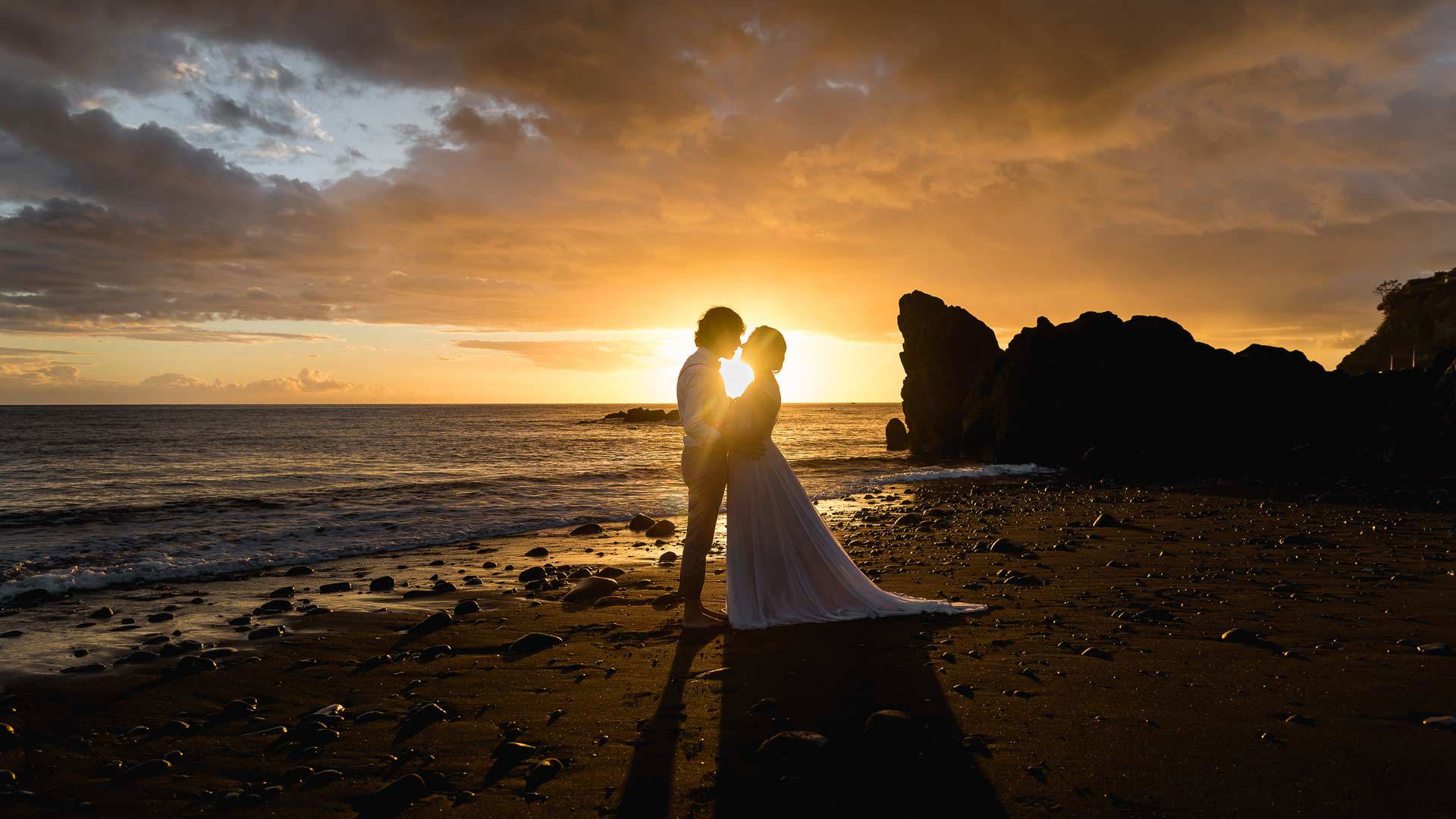 Casal em trajes de casamento a apreciar o pôr do sol na praia.