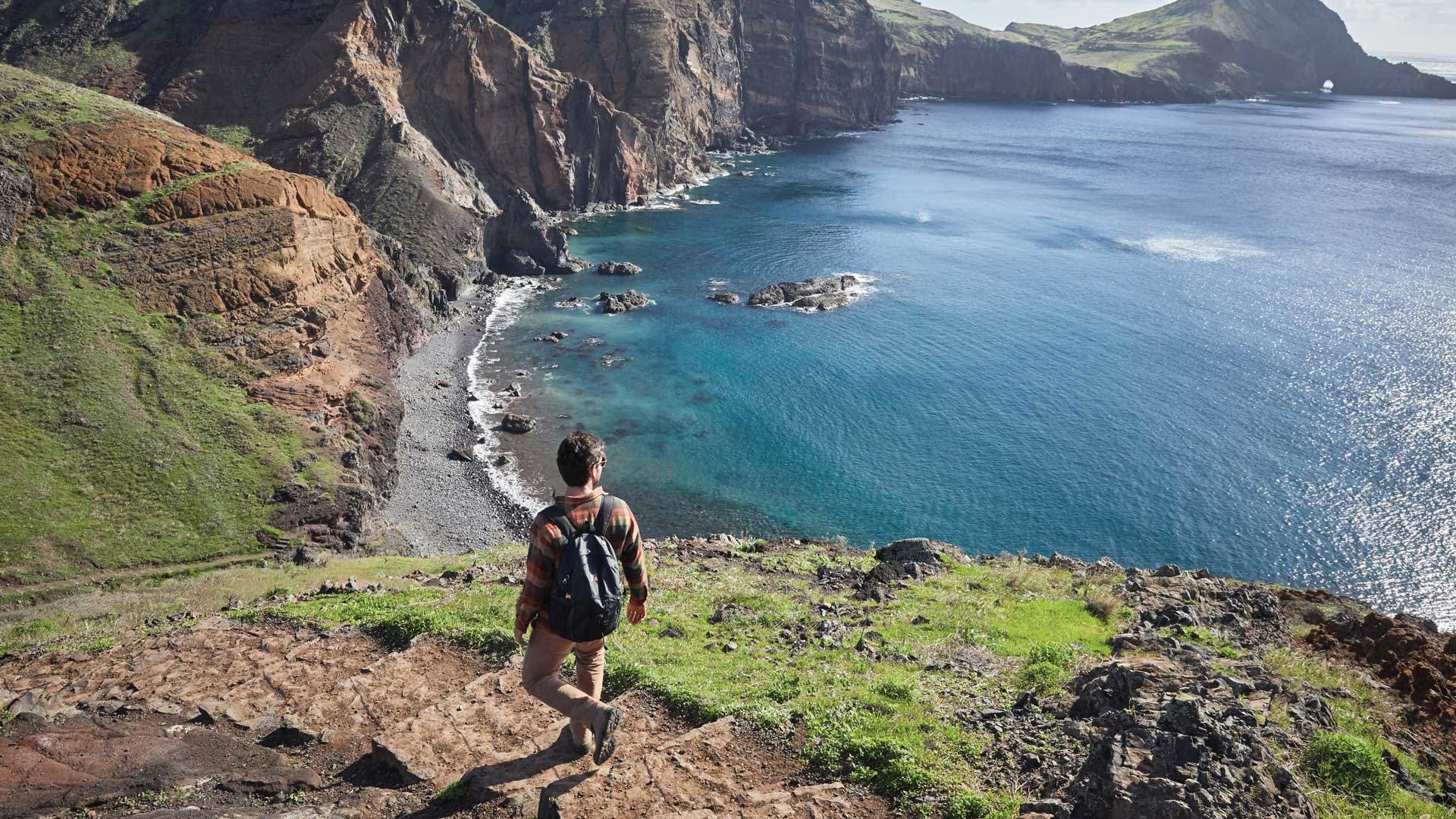 Homem a caminhar por trilho junto à praia de calhau com vista para o mar.