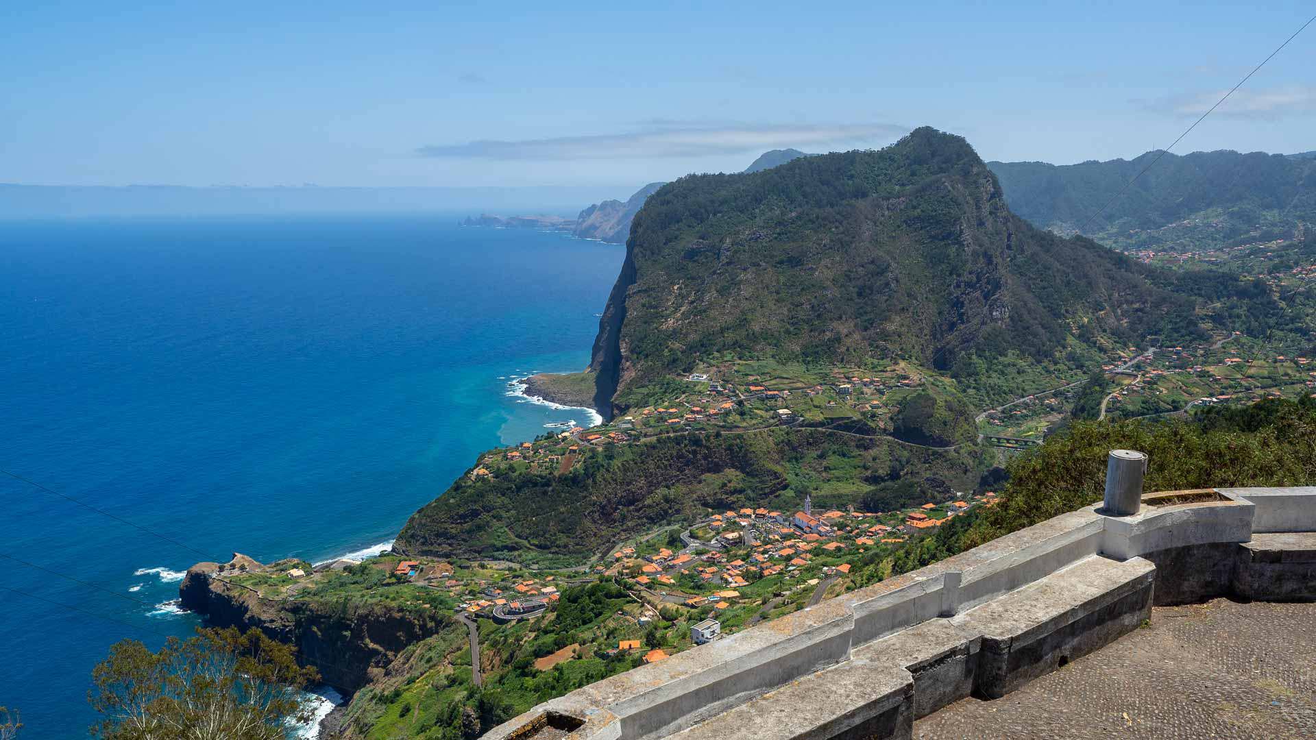 Viewpoint overlooking green valley, houses, and sea in the background.