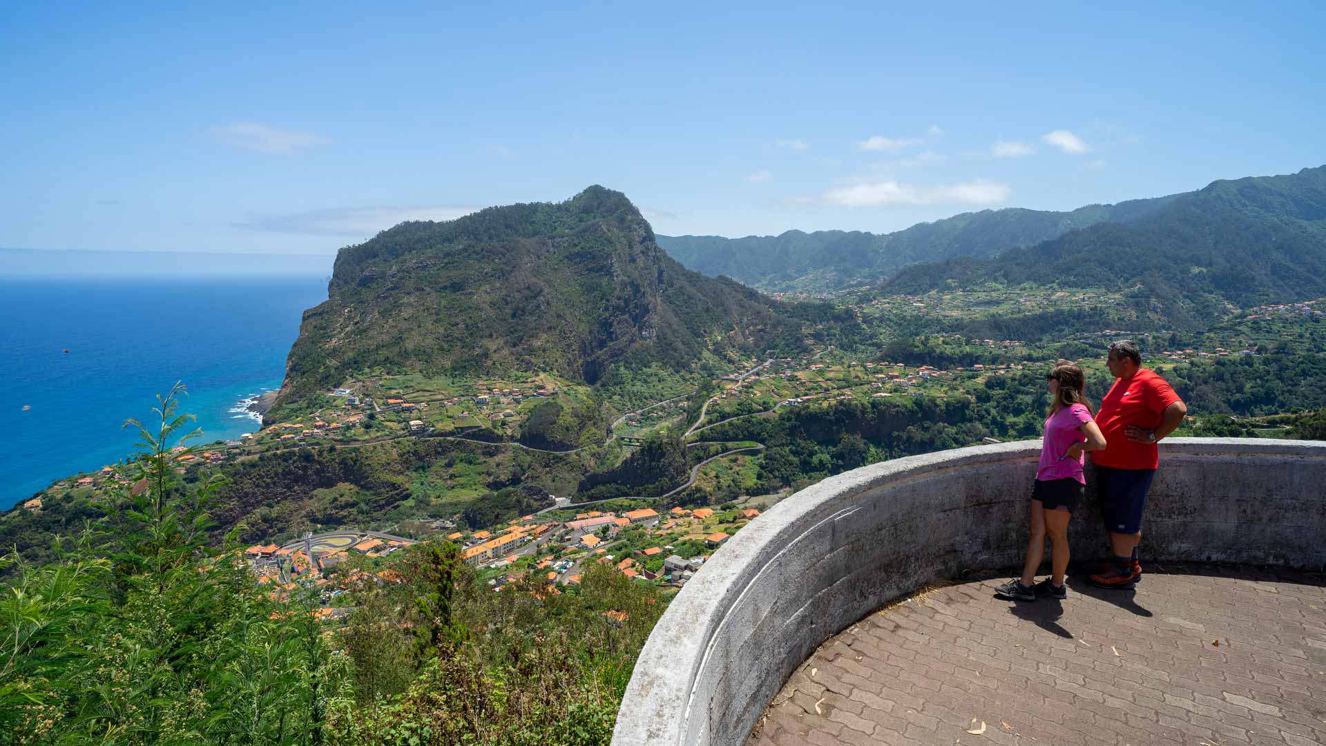 Two people at viewpoint overlooking green valley and houses.