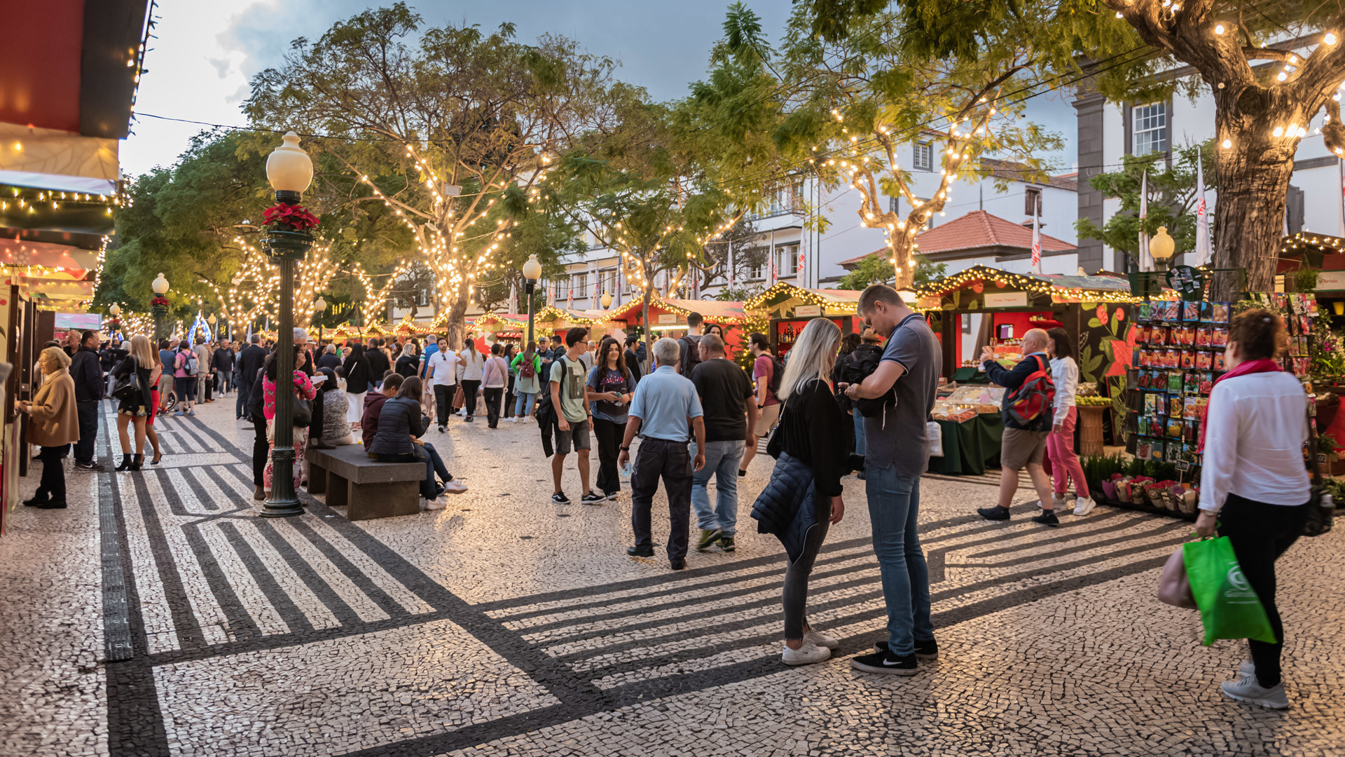 Pessoas junto às barraquinhas de Natal com luzes na placa central do Funchal.