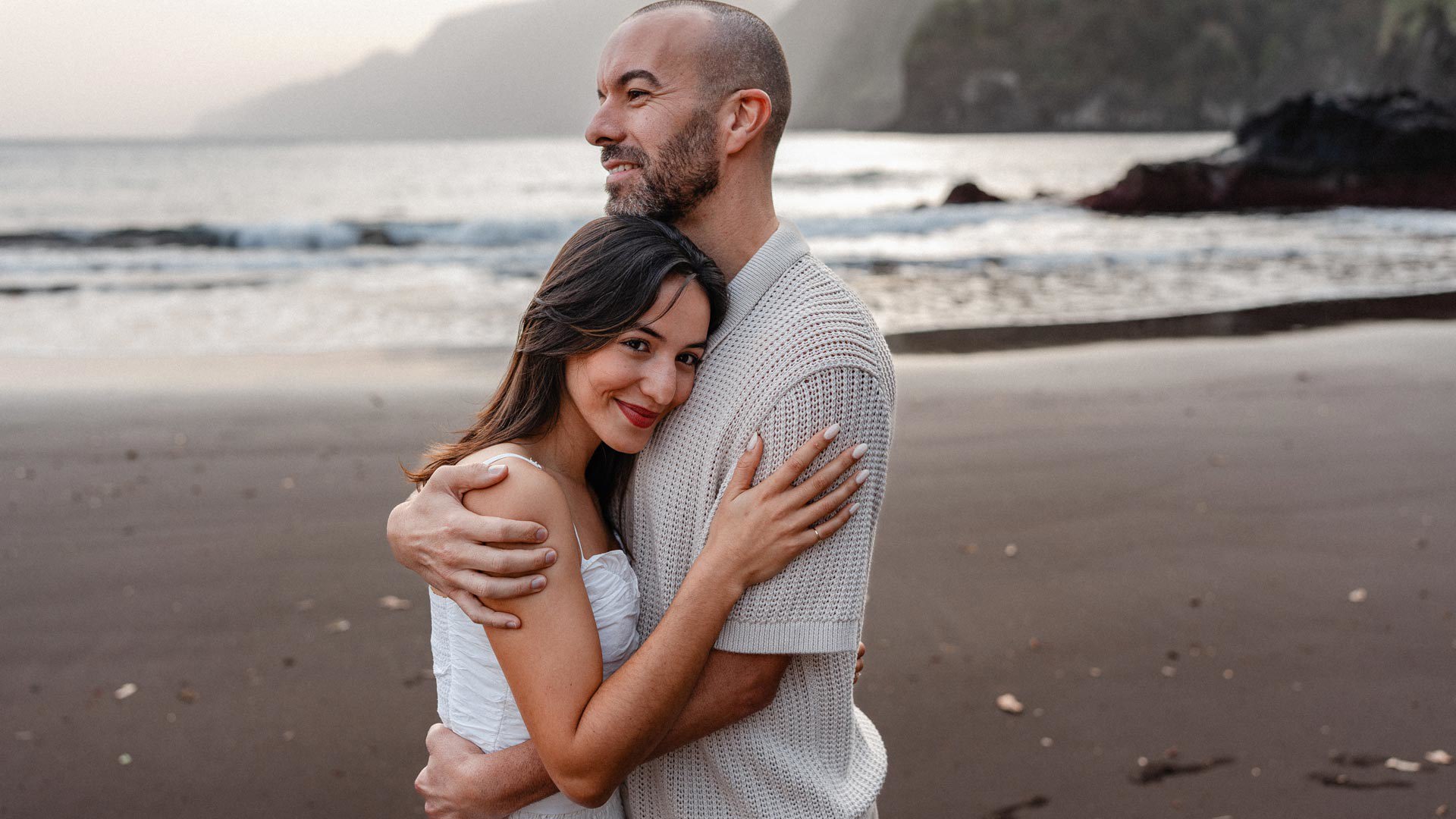 Paar umarmt am schwarzen Sandstrand von Seixal, mit der Küste im Hintergrund.