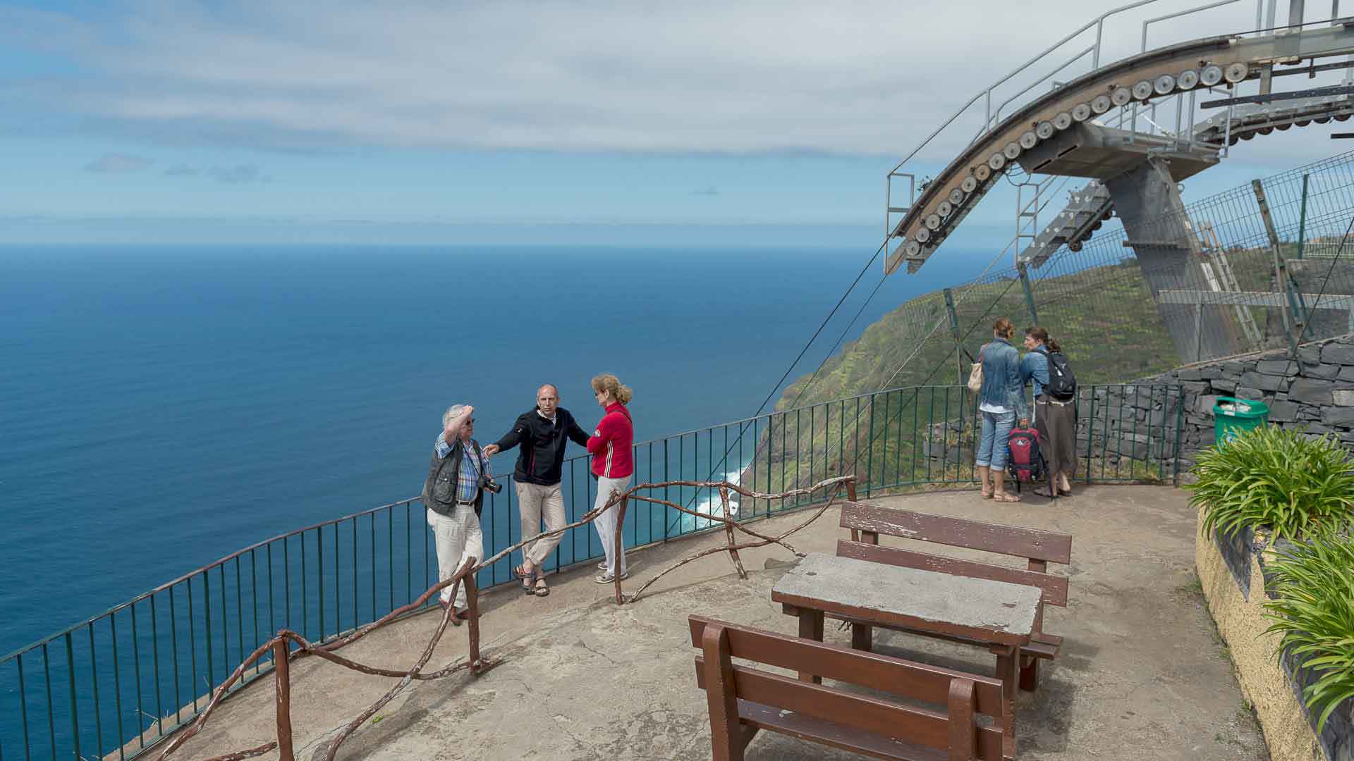 Drei Personen stehen draußen zusammen in der Nähe einer Bank mit Tisch und Meerblick.