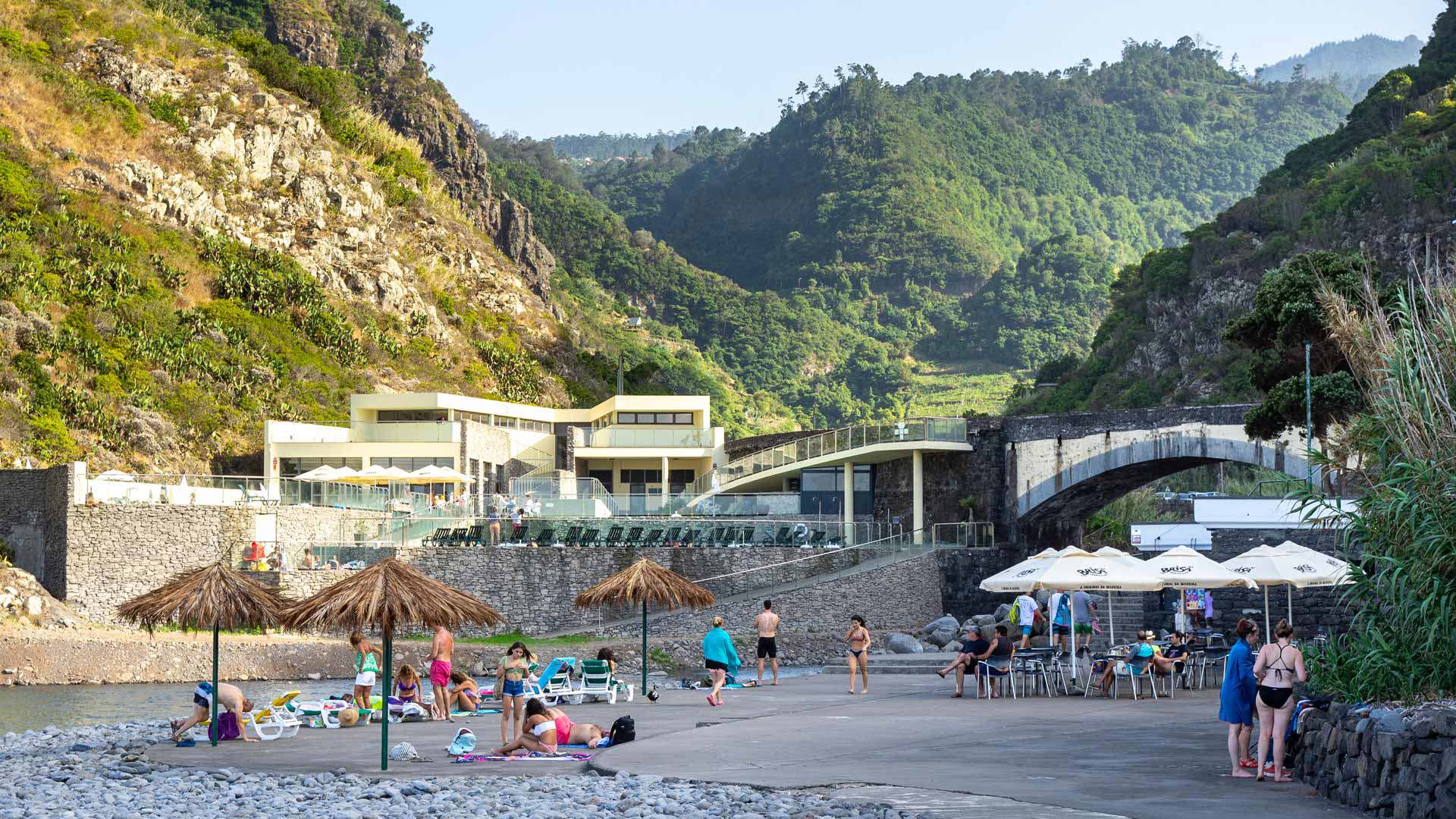 Personas con sombrillas en la playa de guijarros junto a montañas de Madeira.