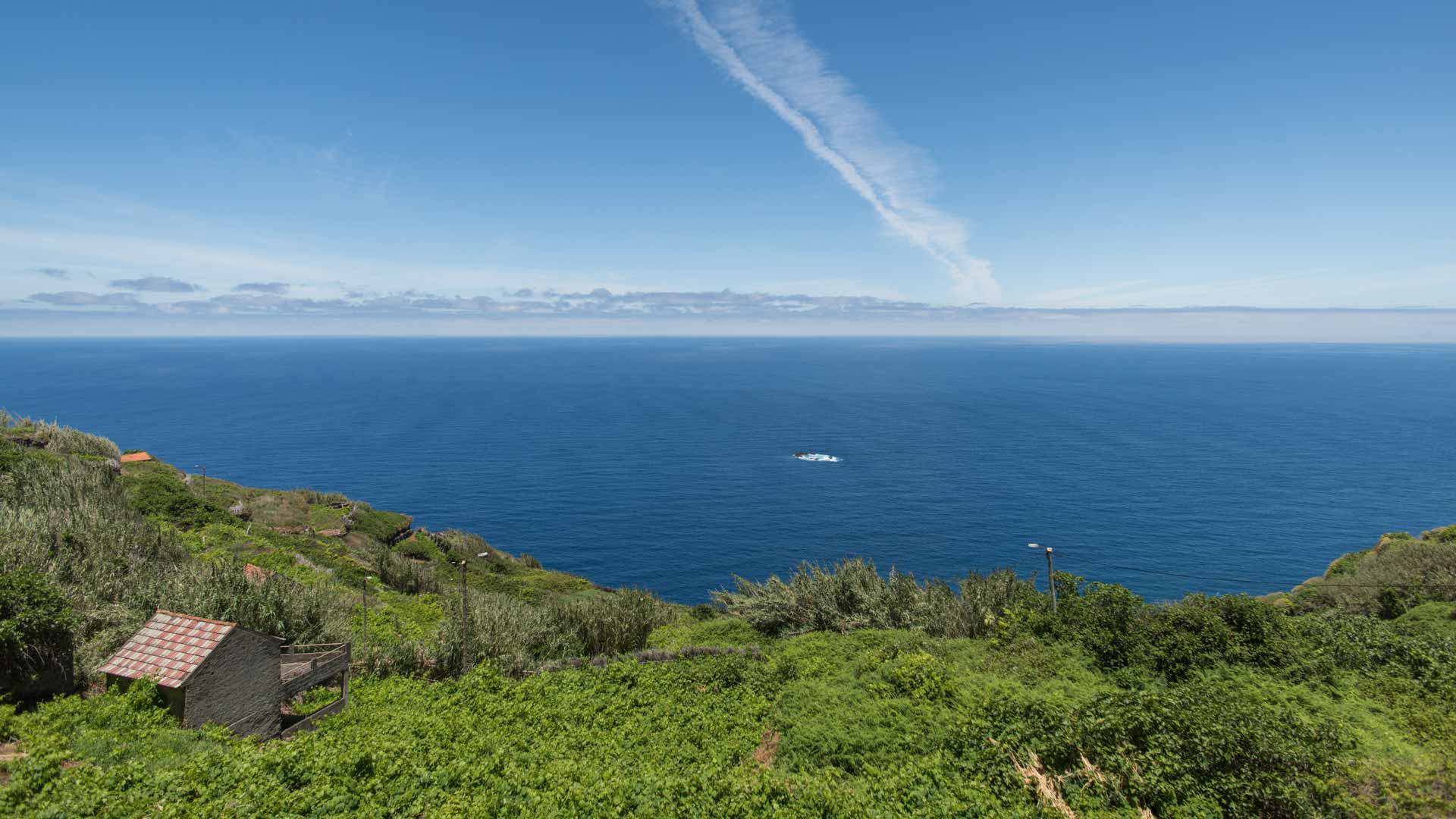Encosta verde com casa de pedra à esquerda e mar ao fundo.