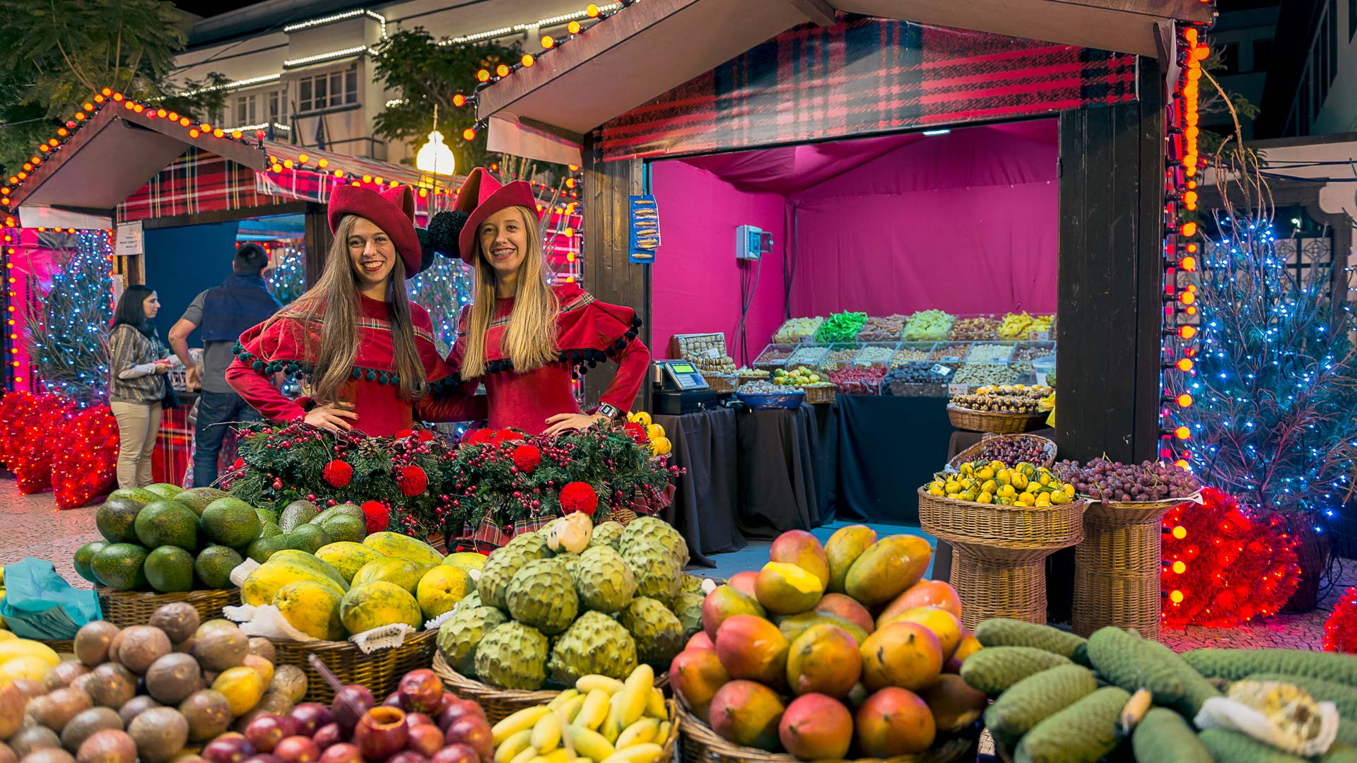 Duas mulheres com trajes de Natal na placa central do Funchal, junto a frutas expostas e barraquinhas.