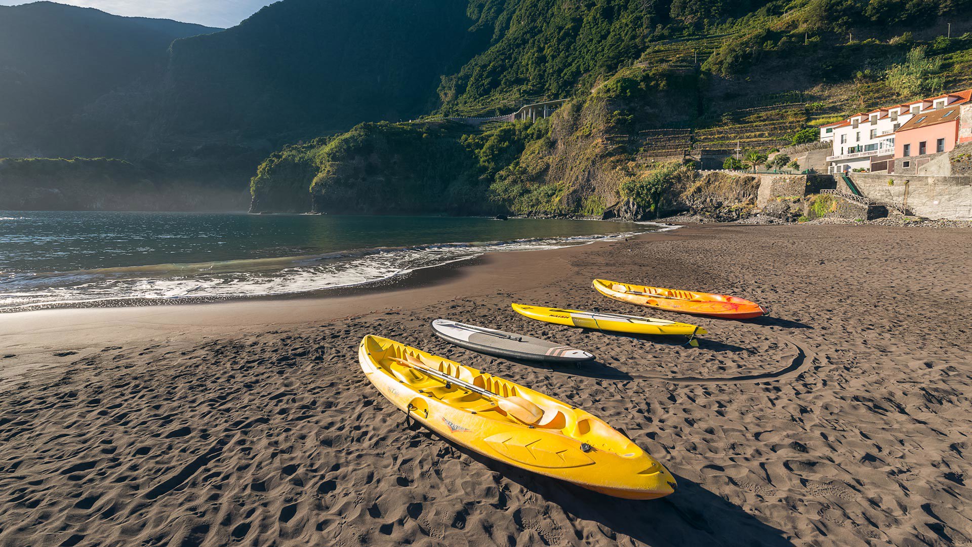 Vier Kajakkanus auf schwarzem Sand am Meer, mit Bergen und Häusern im Hintergrund.