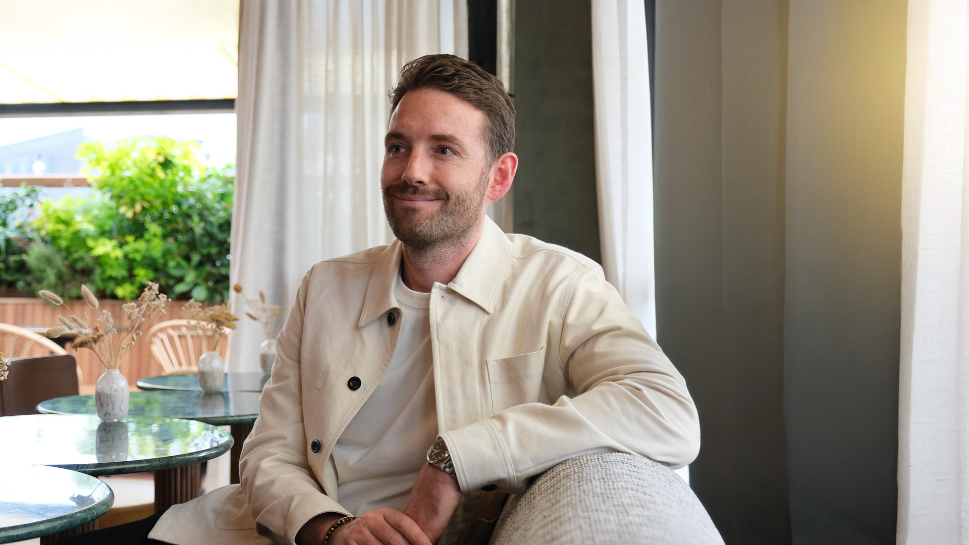 Mann in heller Kleidung lächelt auf einem Sofa sitzend.