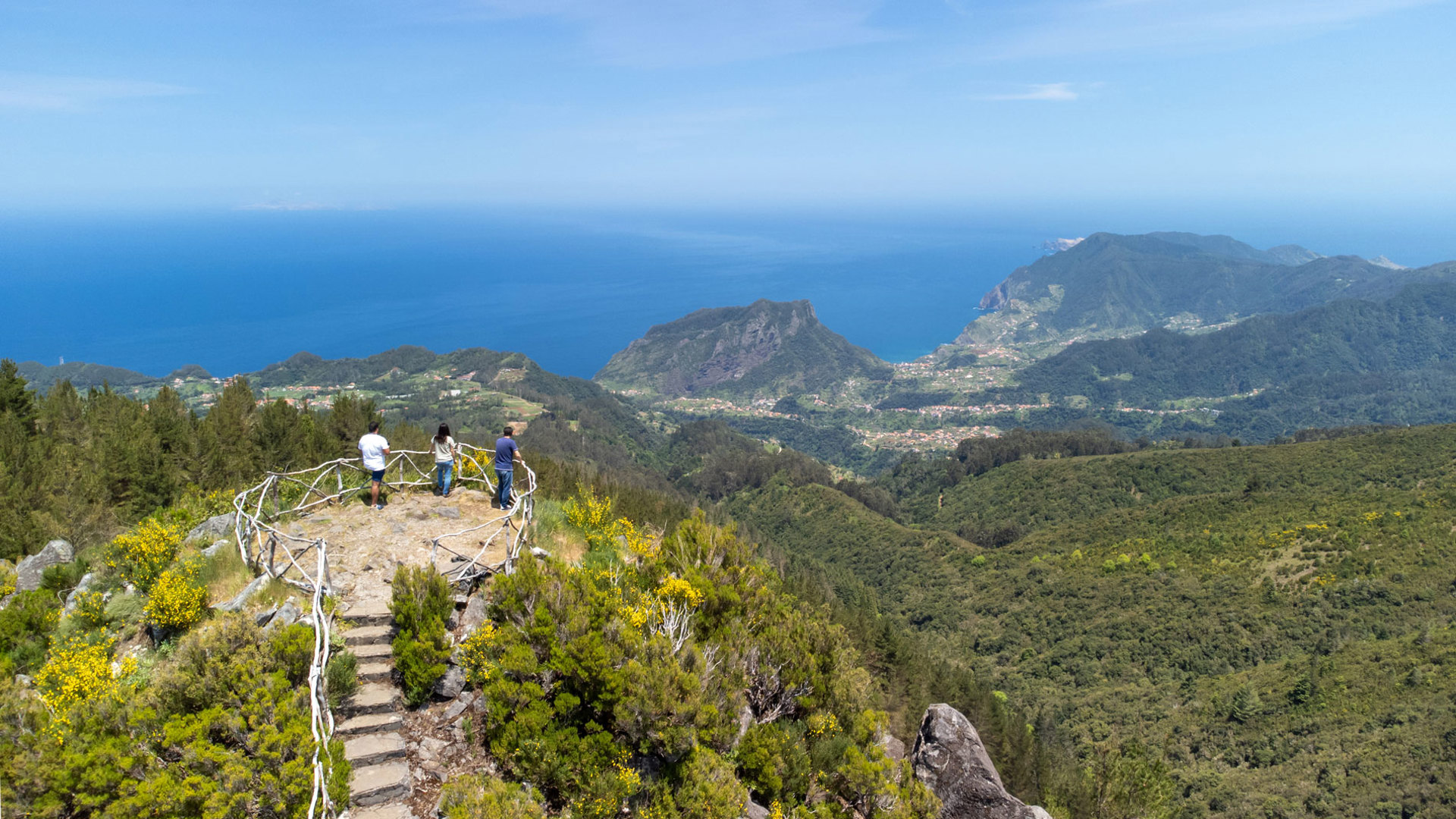 Pessoas num miradouro no meio da vegetação com vista para mar e montanhas.