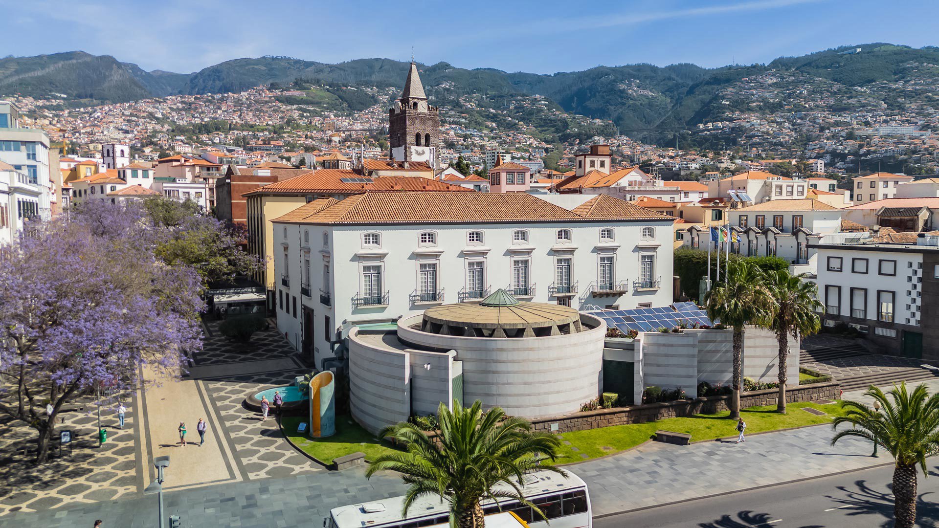 Cidade com edifícios, árvores, estrada e autocarro em movimento.