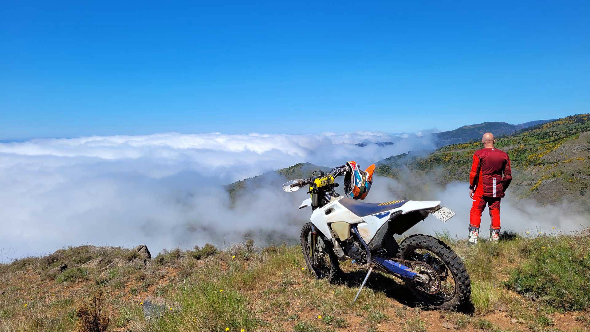 Moto todoterreno parada con persona apreciando la vista bajo nubes, rodeada de naturaleza.