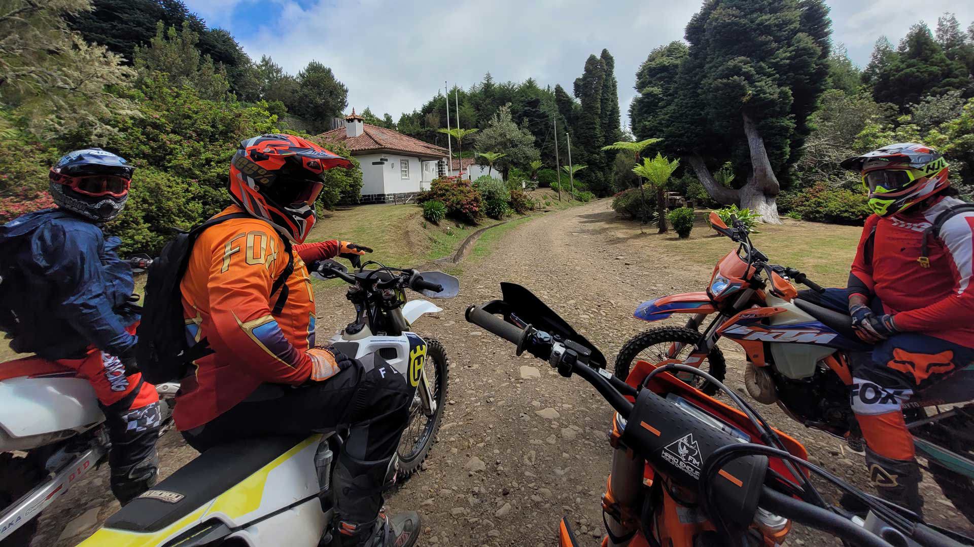 Tres personas en motos de enduro pasando por una casa en terreno de tierra con vegetación.