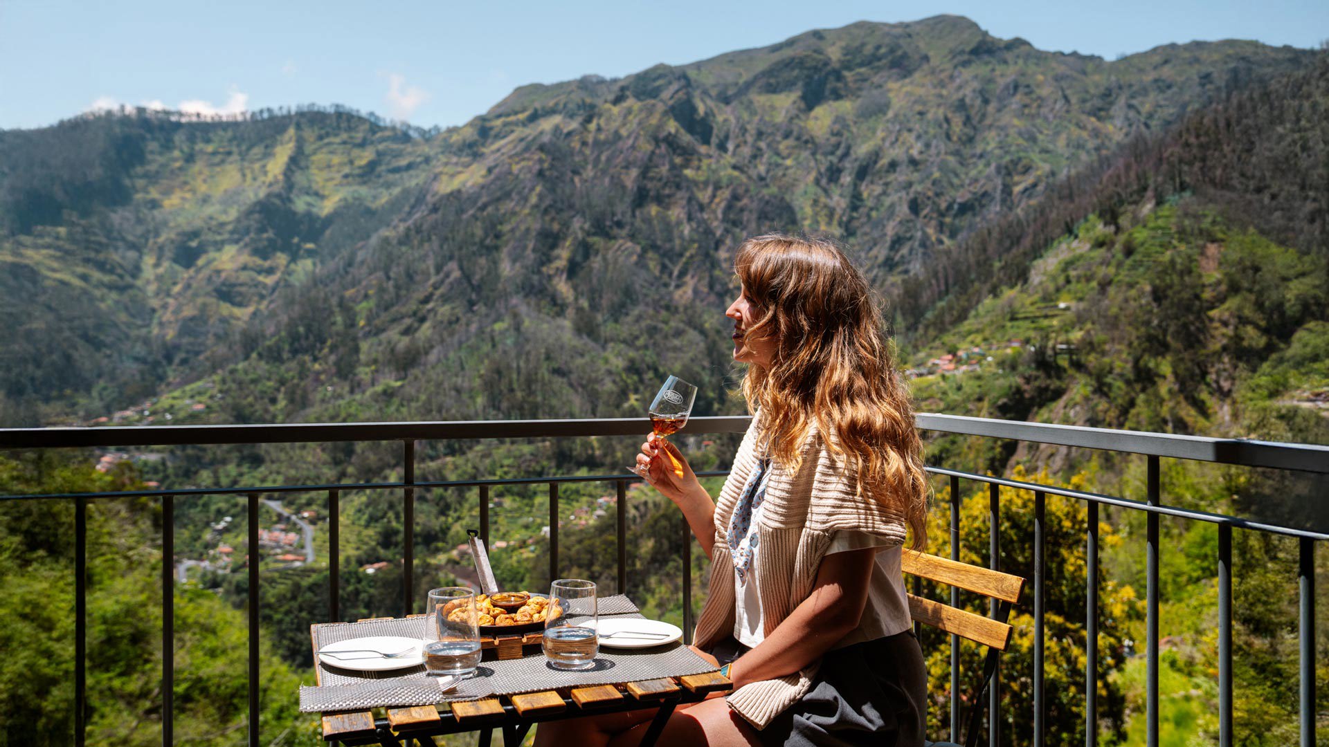 Femme déjeunant sur une terrasse au Curral das Freiras, entourée de montagnes et de végétation.
