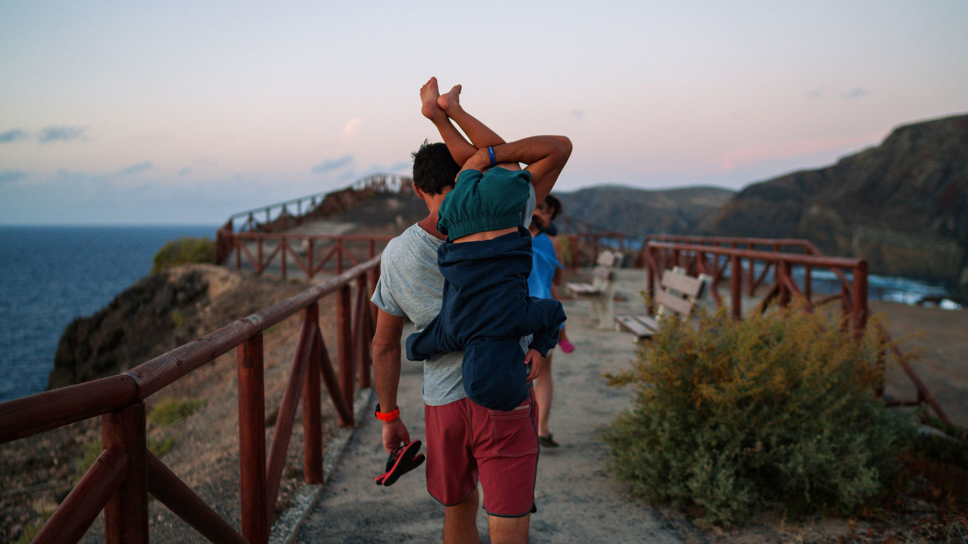 Padre con hijo a caballito en el paseo de la playa en Porto Santo al atardecer, con la ladera al fondo.