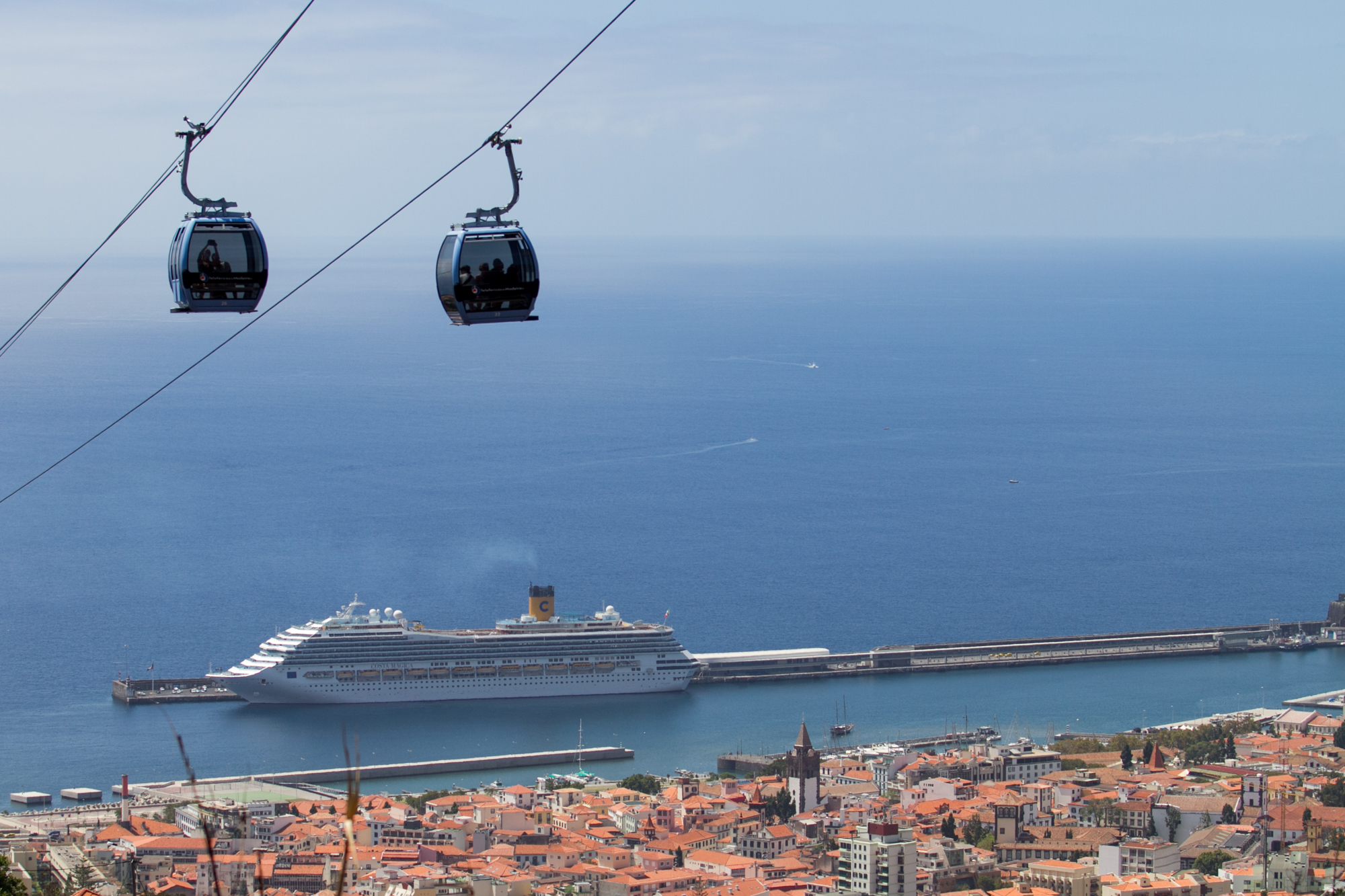 Zwei Seilbahnen über dem Meer mit Kreuzfahrtschiff und Häusern im Hintergrund.