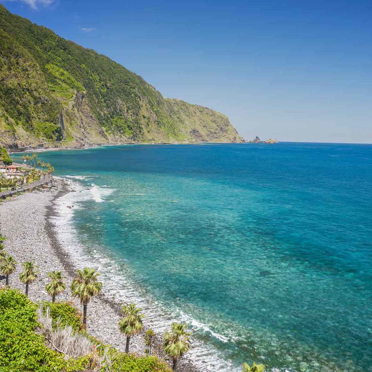 Praia de calhau junto ao mar, com montanha e encosta verde na Madeira.