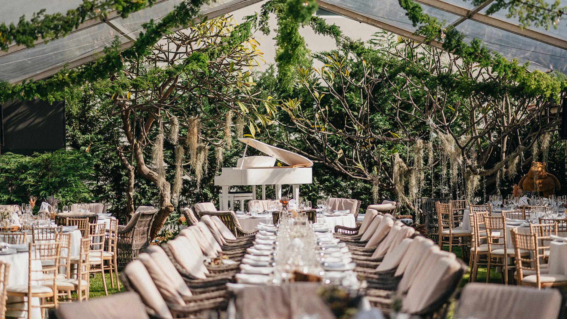 Dining tables decorated under a tent with hanging greenery, piano, and wedding setting.