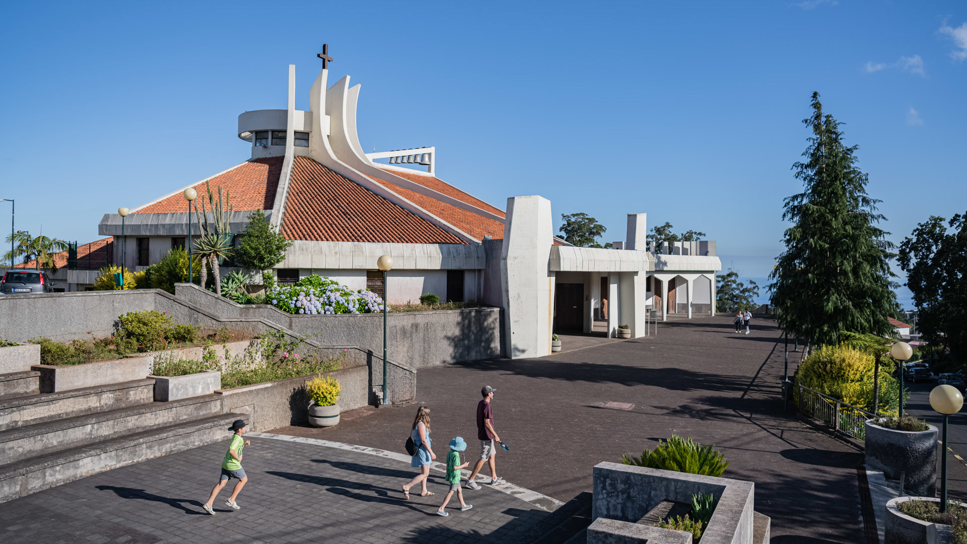 Extérieur d’église avec une famille en promenade et des pots de végétation.