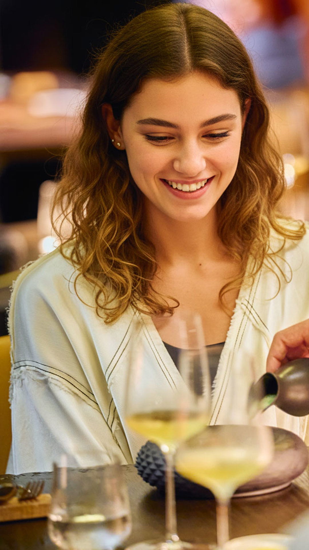 Smiling woman at a table enjoying a meal, with a plate and a glass in front of her.