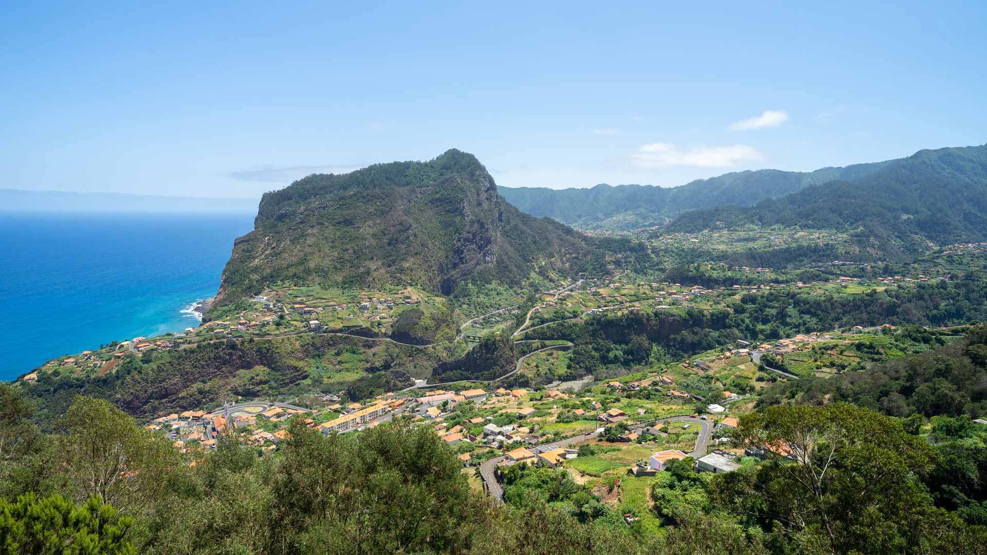 Valle verde con casas entre montañas y vista al mar.
