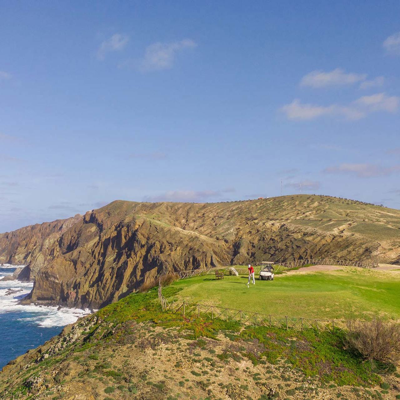Jogador de golfe com buggy num campo em encosta junto ao mar.