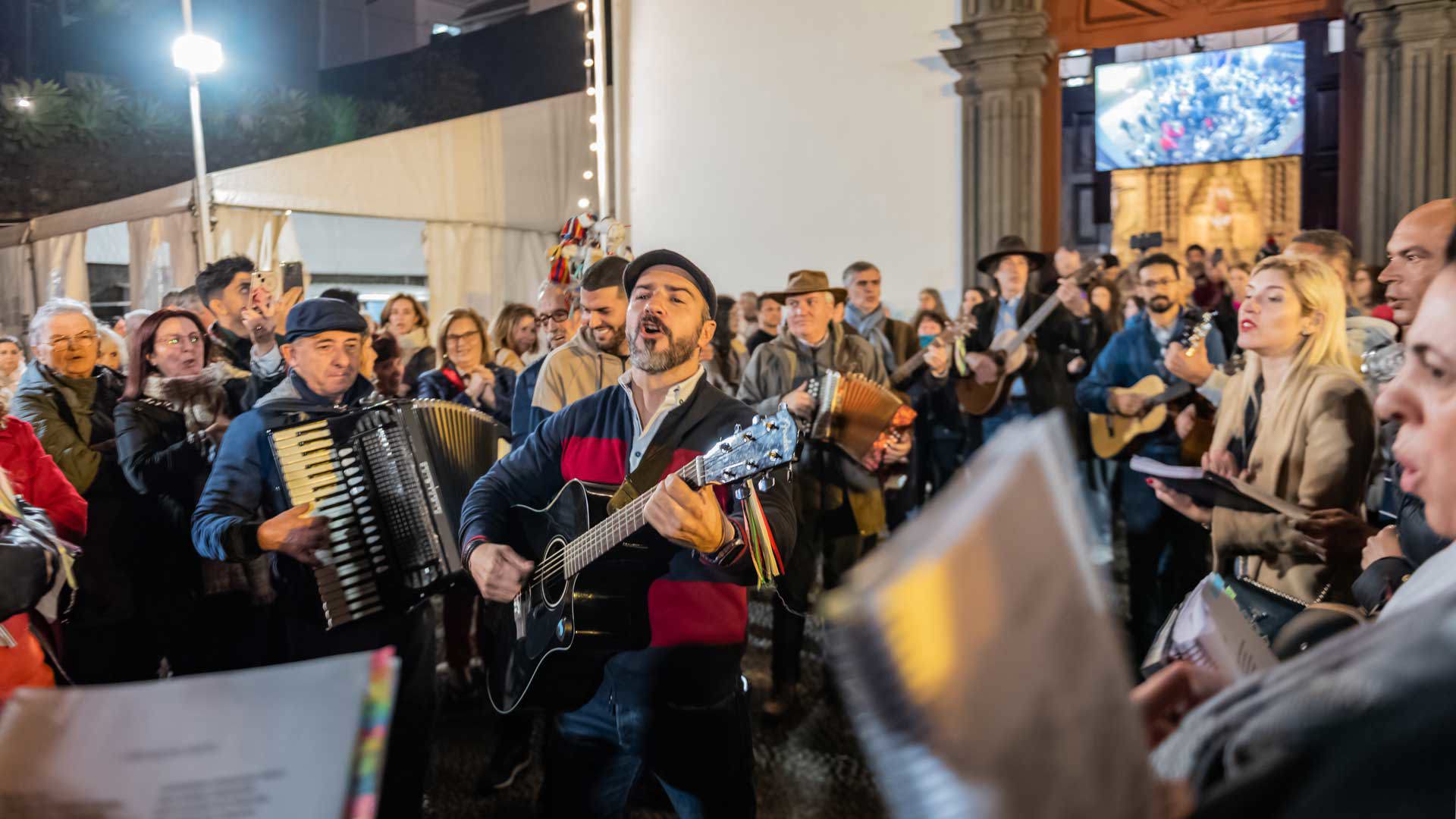 Personas cantando villancicos en las Misas del Parto de Navidad, con instrumentos y iglesia al fondo, Madeira.