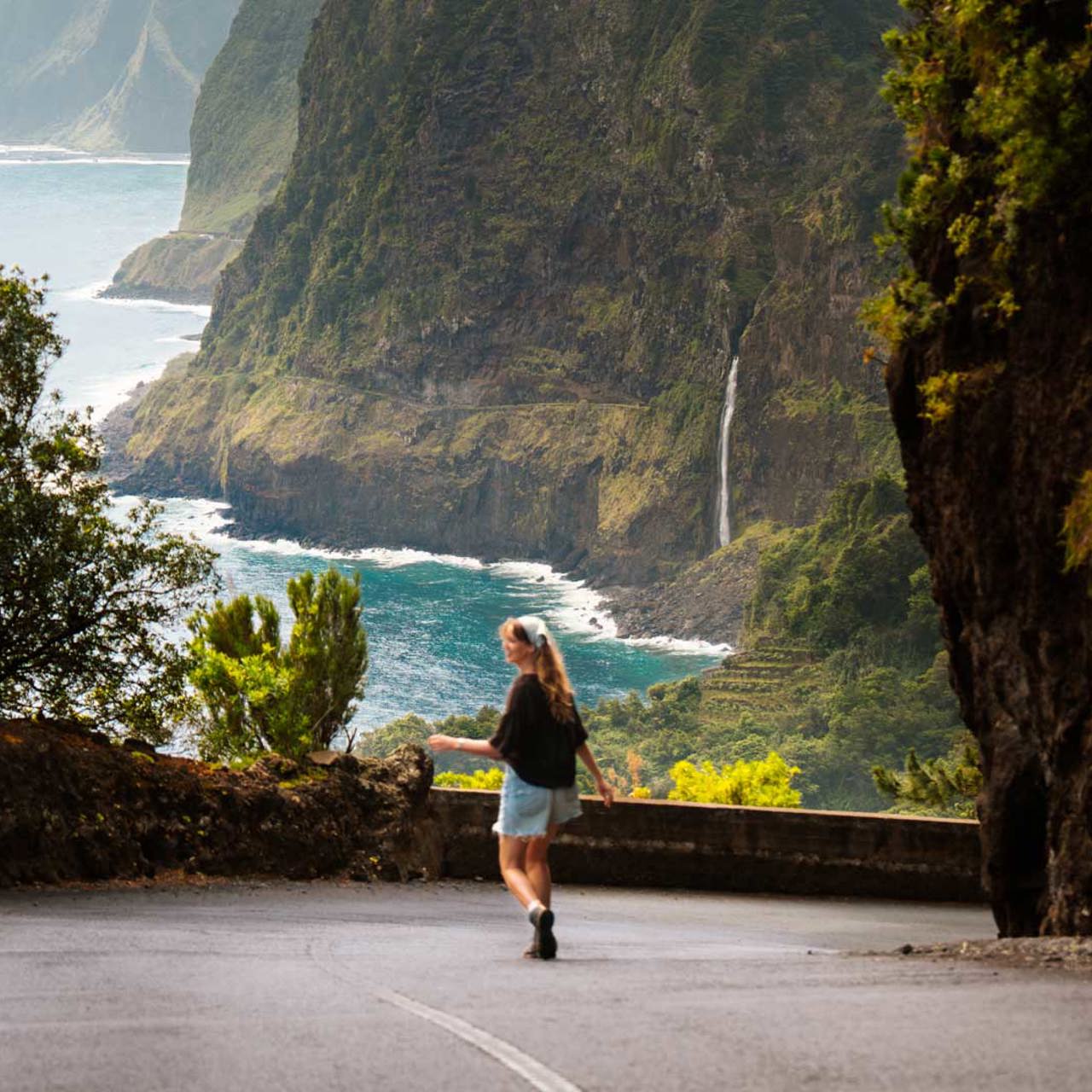 Frau auf einer Straße auf Madeira mit Vegetation, Wasserfall und Meer im Hintergrund.