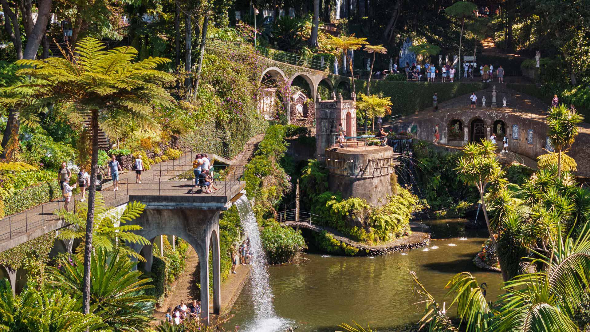 Lago com fontes e pessoas no Jardim Tropical Monte Palace Madeira, rodeado por vegetação.